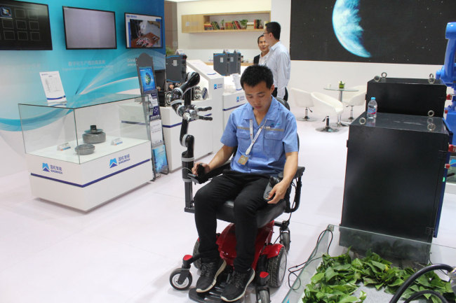 A staff member demonstrates how to operate an intelligent wheelchair with a robotic arm. The wheelchair can be operated by pressing buttons and moving a handle. It can move and fetch things for patients. [Photo: China Plus/Guo Jing]