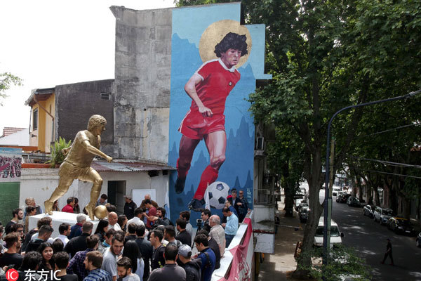 People stand beside statue of Diego Maradona soccer star during its presentation in Buenos Aires, Argentina, Wednesday, October 31, 2018. The statue by artist Jorge Martinez, was presented Wednesday to honor Maradona on his 58th birthday Tuesday, next to the stadium of the Buenos Aires based club where the Argentine soccer great began his career.  [Photo: Imagine China]