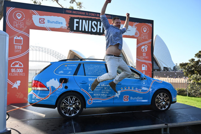 Dutch driver Wiebe Wakker (C) celebrates after driving his retrofitted station wagon nicknamed "The Blue Bandit" onto a platform to complete a round-an-world trip in an electric car with a backdrop of Sydney Harbour Bridge in Sydney on April 7, 2019. Wakker drove some 95,000 kilometres across 33 countries in what he said was the world's longest-ever journey by electric car. [Photo: AFP/Peter Parks]