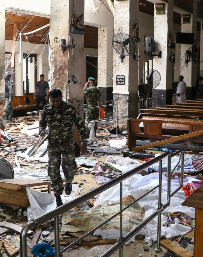Sri Lankan security personnel walk past dead bodies covered with blankets amid blast debris at St. Anthony's Shrine following an explosion in the church in Kochchikade in Colombo on April 21, 2019. [Photo: ISHARA S. KODIKARA / AFP]