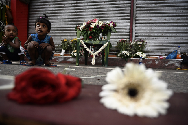 Children sit next to(挨着 āizhe) flowers left by mourners near St. Anthony's Shrine in Colombo on April 23, 2019, two days after a series of bomb blasts targeting(针对 zhēnduì) churches and luxury hotels in Sri Lanka. [Photo: AFP/Jewel SAMAD]